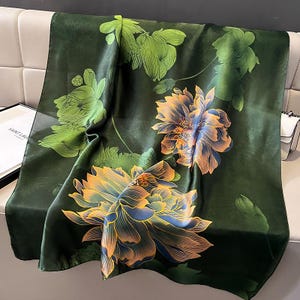 Op de afbeelding: Een donkergroene zijden sjaal met een bloemenpatroon. De sjaal heeft grote, gestileerde bloemen in tinten geel, oranje en blauw, met groene bladeren en stelen. De sjaal is gedrapeerd over een lichtgekleurd oppervlak.