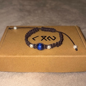 Könnte beinhalten: Ein braunes Makramee-Armband mit einer zentralen blauen Tigeraugen-Perle, flankiert von zwei weißen Perlen. Das Armband hat einen verstellbaren Kordelverschluss mit zwei weiteren weißen Perlen an den Enden. Die Kordel ist in einem Muster geknotet.