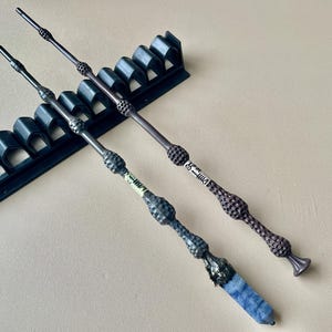 以下が含まれることがあります： 複雑なデザインと暗い色の3つの装飾的な杖が表示されています。 1つの杖は、基部に青い結晶が付いています。 黒い杖ホルダーが背景にあります。 杖の長さは約33cmです。