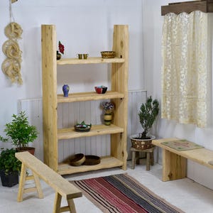 Op de afbeelding: Een rustieke houten boekenplank met vier planken, met diverse decoratieve items zoals vazen en kommen. De natuurlijke houtnerf en het ontwerp met levende rand creëren een warme, uitnodigende uitstraling. Een kleine houten bank en een kleurrijk tapijt zijn ook zichtbaar.