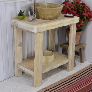 May include: A light-colored wooden vanity with a stone sink and chrome faucet. The vanity has a lower shelf and is supported by four thick, natural-looking legs. A small succulent in a green pot sits on the vanity top.