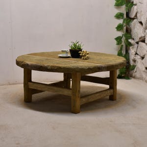 Può includere: Tavolino da caffè rotondo in legno rustico con finitura naturale invecchiata. Il tavolo ha un piano spesso e circolare sostenuto da robuste gambe quadrate. Una piccola pianta in vaso, una tazza da tè e fiori secchi.