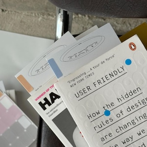Pode incluir: Uma pilha de livros com capas brancas e vários títulos. Um livro é intitulado "USER FRIENDLY" com texto sobre design. Outro livro é intitulado "HA" com o texto "WINNER OF THE". Vários cartões brancos pequenos com um design também são visíveis.