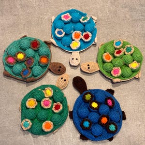 Könnte beinhalten: Fünf gehäkelte Schildkrötenspielzeuge mit bunten Knöpfen auf ihren Panzern. Die Schildkröten sind in verschiedenen Farben: blau, grün, braun und dunkelgrün.