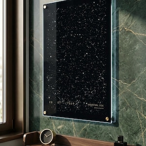 May include: A framed black and white star map with the date 07/20/1969 and the location Houston, USA. The map is mounted with gold-colored hardware on a green marble wall. A small clock and cologne bottle are on a wooden shelf below.