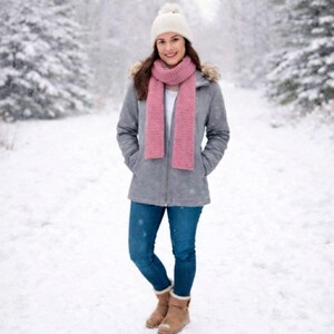 May include: A woman wearing a gray winter coat with a fur-lined hood, a white knit hat with a pom-pom, and a pink scarf. She is wearing blue jeans and brown boots, standing in a snowy forest.