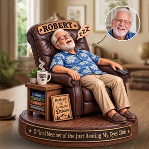 Pode incluir: Uma estatueta personalizada de um homem de cabelos brancos e óculos dormindo numa poltrona de couro castanho. A base contém o texto Official Member of the Just Resting My Eyes Club. Uma mesa lateral tem uma caneca Good Vibes e livros intitulados Travel, Golf, Fishing, Woodworking.
