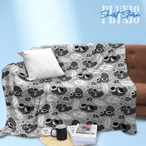 May include: A gray blanket with a repeating cartoon design of a creature with speech bubbles that say "Pebble?" and "There you are!". The blanket is draped over a brown couch with a white pillow.