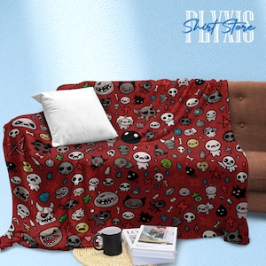 May include: Red throw blanket with a repeating pattern of cartoon characters, skulls, and other graphic elements. A white pillow rests on the blanket, which is draped over a brown sofa. A black mug and books are visible.