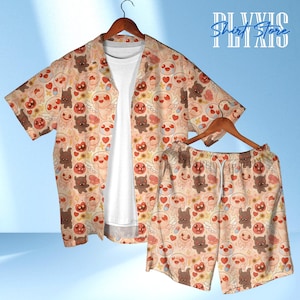 May include: A two-piece matching set featuring a short-sleeved button-down shirt and shorts. The set is beige with a repeating pattern of cartoon characters in brown, pink, and white. The shirt is open, revealing a white undershirt.