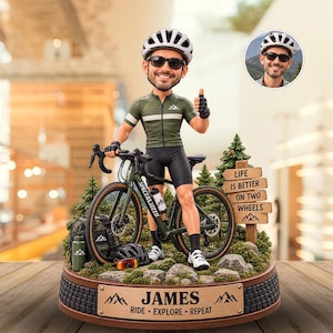 May include: A custom bobblehead figurine of a cyclist wearing a green jersey, black shorts, and a helmet, standing with a bicycle on a base. The base features the name JAMES, the text RIDE EXPLORE REPEAT, and a sign reading LIFE IS BETTER ON TWO WHEELS.
