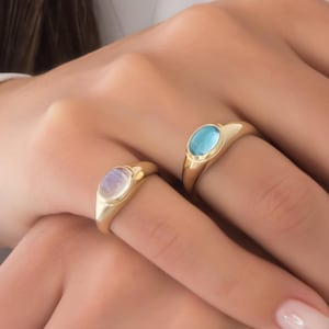 May include: Two gold signet rings, one with a light blue stone and the other with a pearlescent stone, are displayed on a person's fingers. The rings have a classic, elegant design, perfect for everyday wear or special occasions.