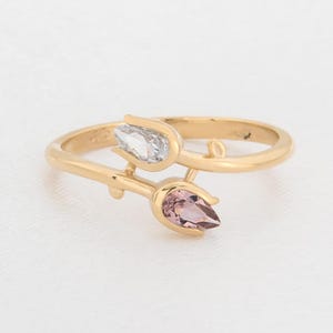 May include: A gold ring with a floral design, featuring two tulip-shaped gemstones. One gemstone is clear, the other is a soft pink. The ring is set against a white background.