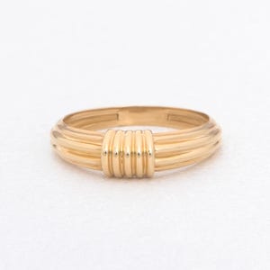 Könnte beinhalten: Ein goldener Ring mit einem einzigartigen Design. Der Ring hat eine Reihe paralleler Bänder, die sich um den Finger wickeln und ein strukturiertes und auffälliges Aussehen erzeugen. Der Ring besteht aus glänzendem, gelbem Gold.