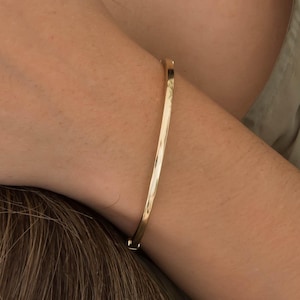 May include: A gold-colored bangle bracelet. The bracelet is thin and has a hammered texture. The bracelet is worn on a wrist. The background is a person wearing a green shirt.