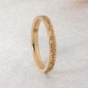Könnte beinhalten: Ein goldfarbener Ring mit floralem Muster. Der Ring ist dünn und filigran, mit einem sich wiederholenden Design aus Blumen und Blättern. Der Ring ist vor einem weißen Hintergrund platziert.