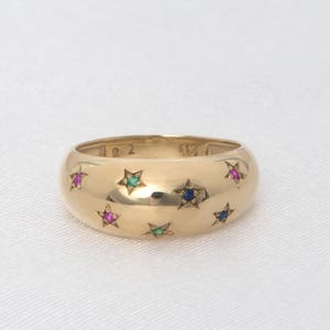 Op de afbeelding: Een gouden ring met een afgeronde band, versierd met kleurrijke stervormige edelstenen. De ring heeft een gepolijst oppervlak en is bezet met roze, groene en blauwe edelstenen. De ring is een sieraad.