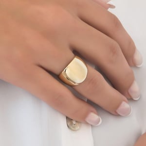 May include: A gold-colored signet ring with a smooth, rounded face. The ring is worn on a finger, set against a white shirt. The ring's design is simple and classic, reflecting a minimalist aesthetic. The ring is a statement piece.