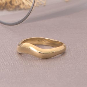 Può includere: Un anello dorato dal design ondulato. L'anello è posto su uno sfondo neutro, con un bicchiere e fiori secchi sullo sfondo. La superficie dell'anello riflette la luce, evidenziando la sua consistenza liscia.