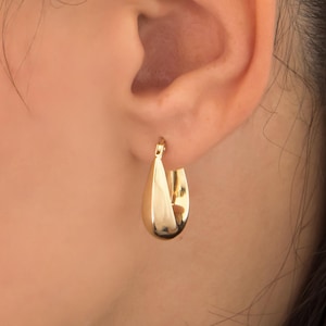 May include: A gold-colored, teardrop-shaped hoop earring. The earring is worn on the ear, and the smooth, polished surface reflects light. The earring has a simple, elegant design.