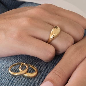 May include: A close-up of three gold rings. One is a large, teardrop-shaped signet ring. Two smaller, simple gold bands are also visible. The rings are on a denim background.