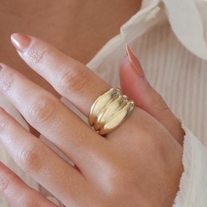 May include: A gold ring with a layered, petal-like design. The ring is worn on a finger, and the hand is resting against a white collared shirt. The ring's surface has a smooth, polished finish, reflecting light.