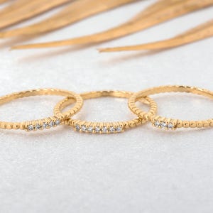 Puede incluir: Tres anillos dorados con pequeñas piedras preciosas transparentes dispuestas en fila. Los anillos tienen una banda texturizada con pequeños detalles redondos. Los anillos se muestran sobre una superficie blanca, con un fondo borroso de hojas doradas.