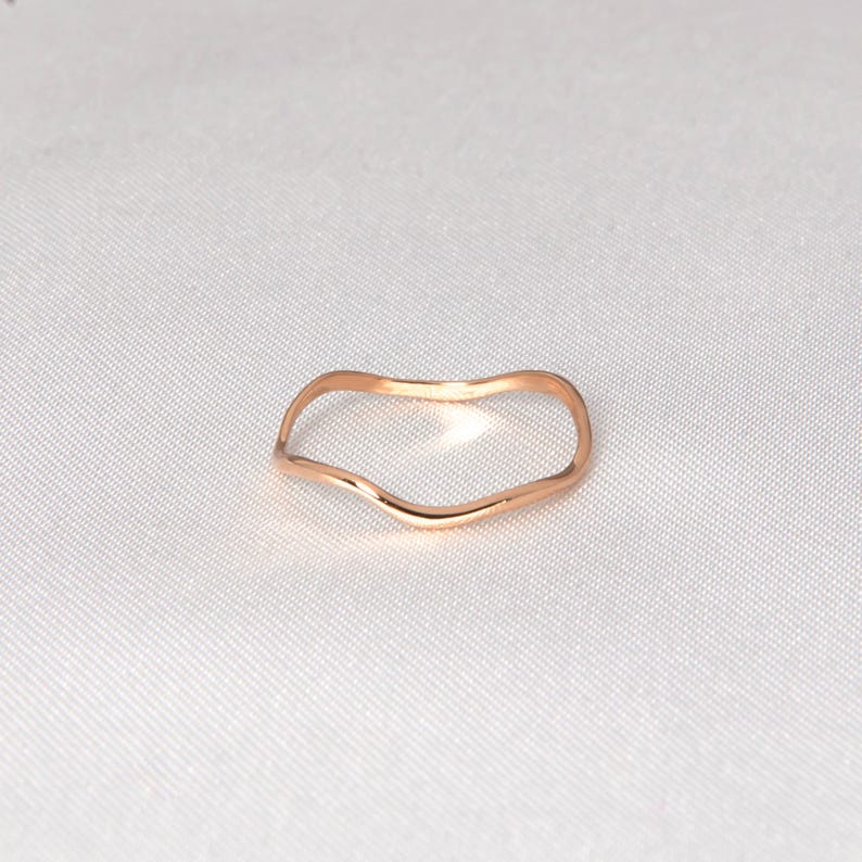 Puede incluir: Un anillo de oro rosa con una forma ondulada y org&aacute;nica. El anillo se presenta sobre un fondo blanco, resaltando su superficie lisa y pulida. El dise&ntilde;o minimalista del anillo lo convierte en una joya vers&aacute;til.