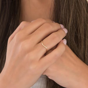 Puede incluir: Un delicado anillo dorado con un dise&ntilde;o ondulado. El anillo es delgado y minimalista, con una suave curva a trav&eacute;s del dedo. El anillo se muestra en el dedo de una persona, destacando su elegancia. Una joya.