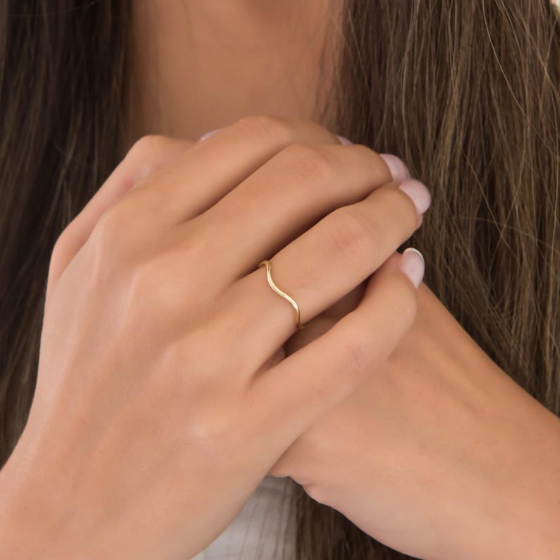 Puede incluir: Un delicado anillo dorado con un dise&ntilde;o ondulado. El anillo es delgado y minimalista, con una suave curva a trav&eacute;s del dedo. El anillo se muestra en el dedo de una persona, destacando su elegancia. Una joya.