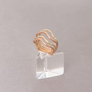 Puede incluir: Un anillo multicolor con un dise&ntilde;o ondulado. El anillo presenta tres bandas en oro, oro rosa y oro amarillo. El anillo se exhibe sobre un soporte cuadrado transparente sobre un fondo neutro. El dise&ntilde;o del anillo es moderno y elegante.