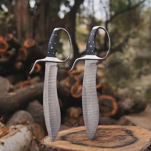 May include: Two handmade knives with curved blades and black handles with silver accents. The knives have a unique design with a loop guard and are displayed on a wooden surface, with a blurred background of logs and foliage.