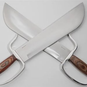 May include: Two crossed silver-bladed knives with wooden handles. The blades are wide and curved, and the handles feature a finger loop. The wood grain on the handles is visible. The knives are displayed against a white background.
