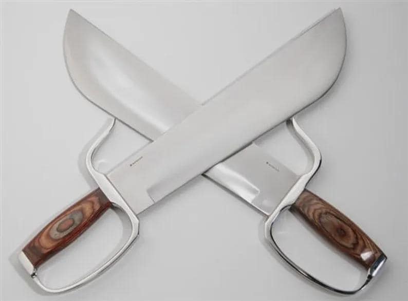 May include: Two crossed silver-bladed knives with wooden handles. The blades are wide and curved, and the handles feature a finger loop. The wood grain on the handles is visible. The knives are displayed against a white background.