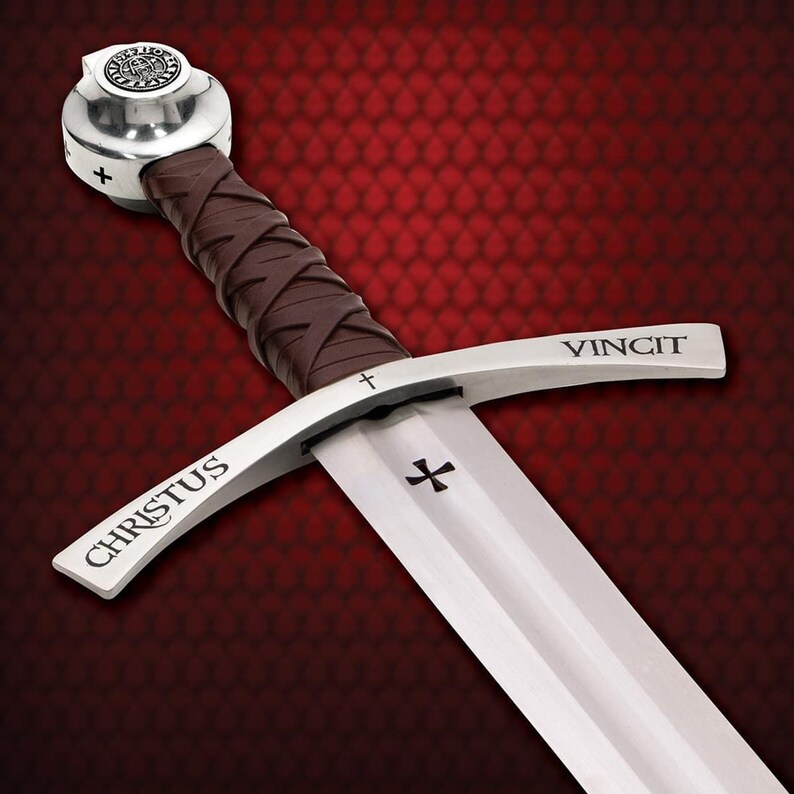 May include: A medieval sword with a silver blade and guard, featuring the inscription "CHRISTUS" and "VINCIT". The handle is wrapped in brown leather. The pommel is silver with a circular emblem and a cross.