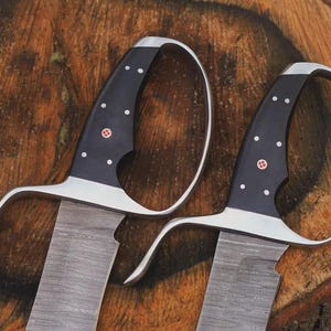 May include: Two swords with black handles, silver guards, and textured blades. The handles have white dot accents and a red emblem. The blades have a wood grain pattern. The swords are displayed on a wooden surface.