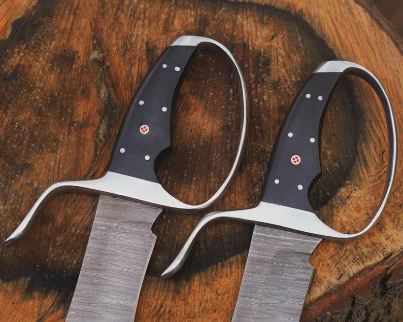 May include: Two swords with black handles, silver guards, and textured blades. The handles have white dot accents and a red emblem. The blades have a wood grain pattern. The swords are displayed on a wooden surface.