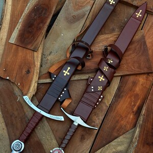 May include: Two swords with brown leather handles and sheaths, adorned with gold crosses. The blades are silver, and the hilts feature decorative elements. One sword has the word "VINCIT" and the other has "CHRISTUS" engraved on the blade.