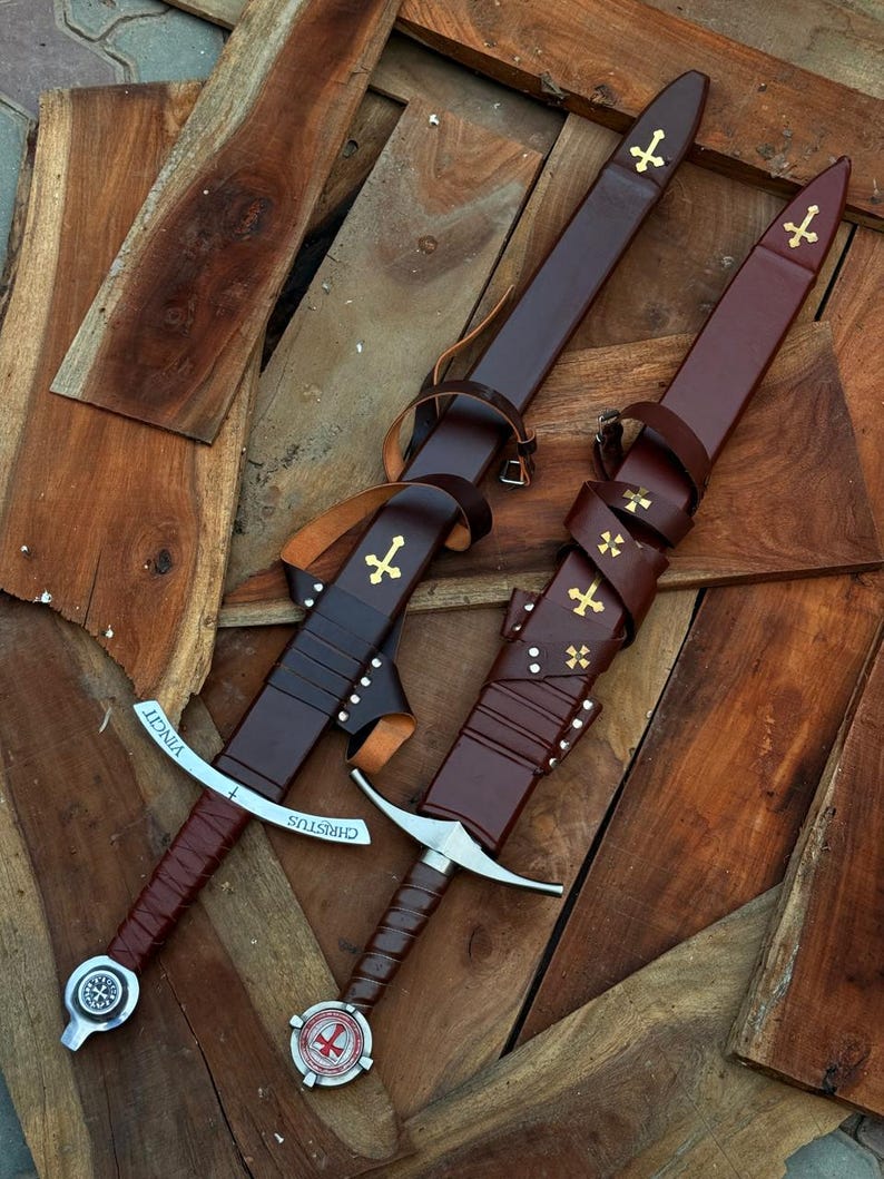 May include: Two swords with brown leather handles and sheaths, adorned with gold crosses. The blades are silver, and the hilts feature decorative elements. One sword has the word "VINCIT" and the other has "CHRISTUS" engraved on the blade.
