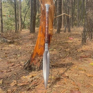 May include: A spear with a silver blade and a long, brown wooden shaft. The shaft is wrapped with a spiral of leather or a similar material. The spear is standing upright in a forest setting, leaning against a tree.