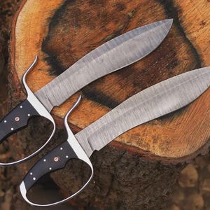 May include: Two large, ornate knives with curved blades and black handles with white dot accents. The blades have a textured pattern. The knives are resting on a wooden surface, showcasing their craftsmanship and design.