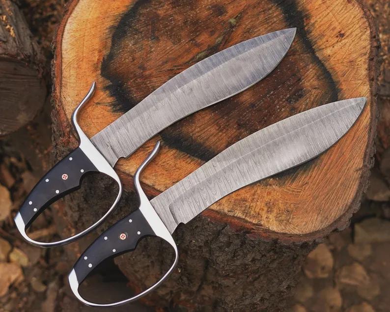 May include: Two large, ornate knives with curved blades and black handles with white dot accents. The blades have a textured pattern. The knives are resting on a wooden surface, showcasing their craftsmanship and design.