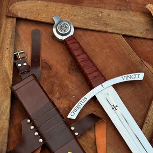 May include: A medieval-style sword with a brown leather-wrapped handle and a silver blade. The blade has the word "CHRISTUS" and "VINCIT" etched on it. A brown leather sheath with a gold cross is also present.