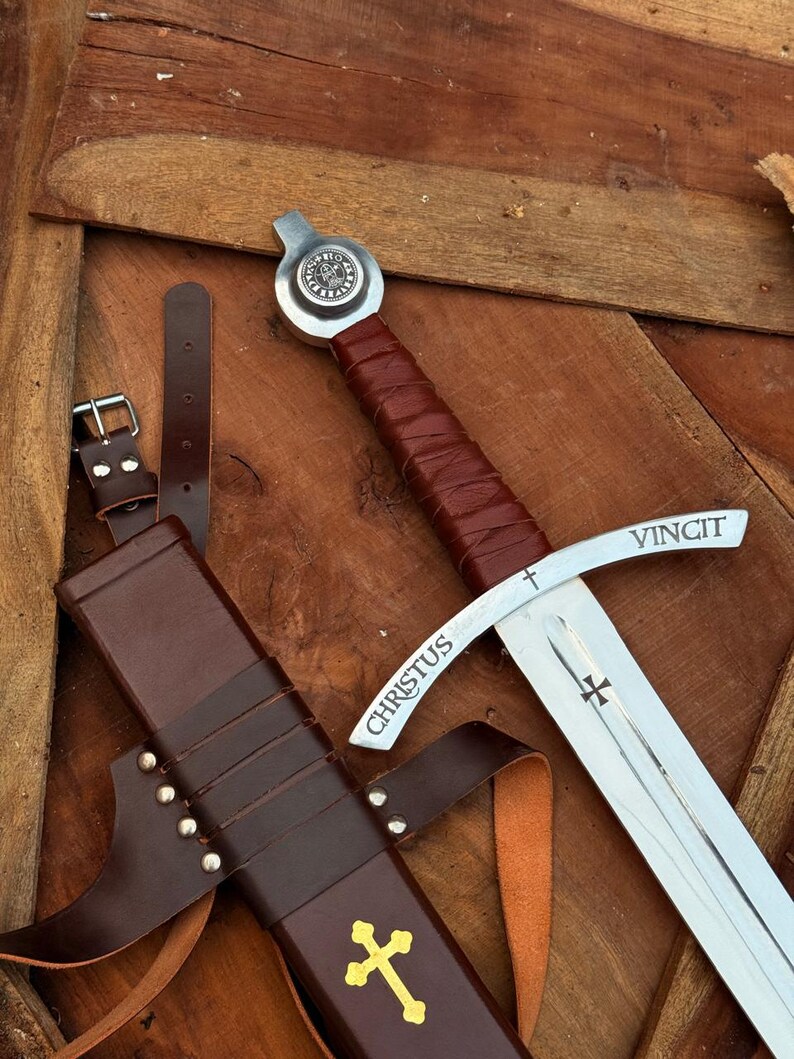 May include: A medieval-style sword with a brown leather-wrapped handle and a silver blade. The blade has the word "CHRISTUS" and "VINCIT" etched on it. A brown leather sheath with a gold cross is also present.