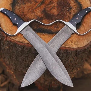 May include: Two crossed, hand-forged knives with curved handles and wide blades. The handles are black with silver accents and small white dots. The blades have a textured pattern, and the knives are set against a wooden background.