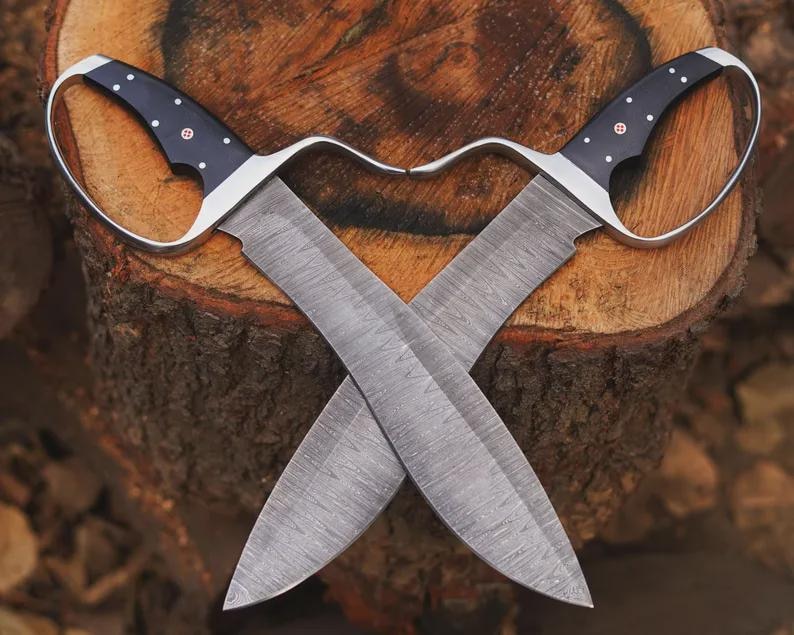May include: Two crossed, hand-forged knives with curved handles and wide blades. The handles are black with silver accents and small white dots. The blades have a textured pattern, and the knives are set against a wooden background.