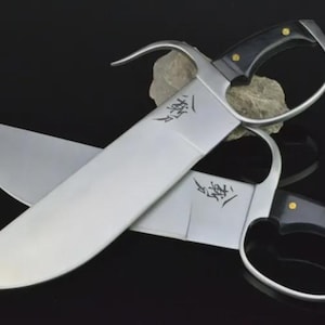 Pu&ograve; includere: Due spade con lame argentate, manici neri e anelli per le dita. Le lame hanno una forma curva e un carattere cinese inciso. I manici sono neri con rivetti dorati. Le spade sono esposte su una superficie scura.