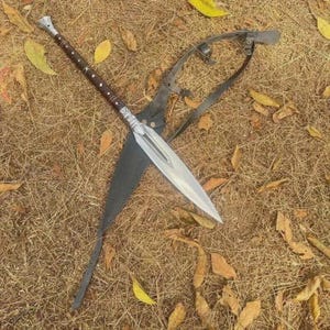 May include: A spear with a silver blade and a long, brown handle with silver accents. The spear is resting on a bed of dry grass and leaves, with a black leather sheath and a metal harness.