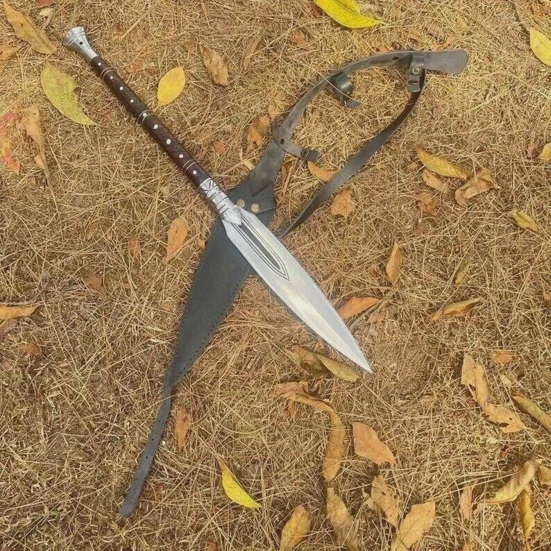 May include: A spear with a silver blade and a long, brown handle with silver accents. The spear is resting on a bed of dry grass and leaves, with a black leather sheath and a metal harness.