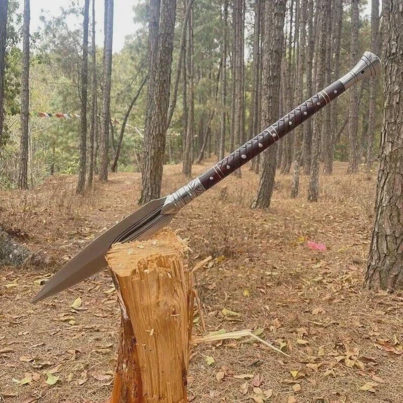 May include: A spear with a long, sharp, silver blade and a dark brown wooden shaft. The shaft is decorated with silver bands and small, dark-colored accents. The spear is resting on a wooden stump in a forest setting.